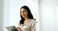 Young woman with long dark hair and olive complexion wearing a beige button-up shirt, holding and viewing a tablet device in a bright, minimalist white interior space.