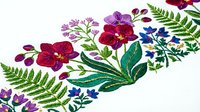 Colorful embroidered floral border with purple and red flowers, green ferns and leaves on white fabric. Decorative textile design for crafts.