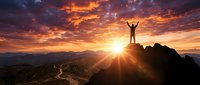 Silhouette of successful person with raised arms standing on mountain peak at dramatic sunset with golden sky and valley landscape below.