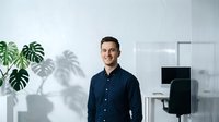 Young professional businessman in dark shirt smiling confidently in modern office workspace with plants and computer setup for corporate presentations.