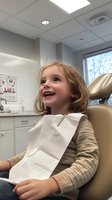 Cheerful young patient with blonde hair smiling during dental checkup, wearing beige sweater in modern medical office with examination chair and anatomical charts.