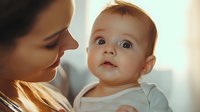 Young Caucasian mother holding baby with bright blue eyes in white onesie, sharing tender moment in soft natural window light, expressing maternal love and bonding.