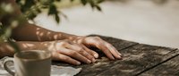 Woman hands resting on rustic wooden table with coffee cup in natural sunlight creating peaceful morning atmosphere for relaxation and mindfulness concepts.