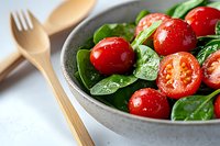 Fresh spinach salad with cherry tomatoes in ceramic bowl, healthy vegetarian meal with wooden utensils on marble surface for cooking and nutrition content.