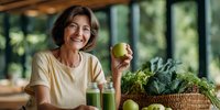 Smiling middle-aged Caucasian woman holding green apple with fresh vegetables and green smoothies in modern kitchen promoting healthy lifestyle and nutrition.