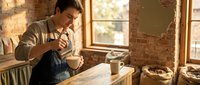 Young male craftsman working with wood in rustic studio workshop, sanding or finishing wooden bowl with hand tools, natural light from windows, artisan furniture making and woodworking craft.