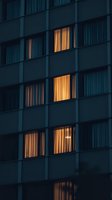 Illuminated apartment windows glowing warmly against dark building facade at dusk, creating mysterious urban atmosphere with curtained windows and geometric patterns.