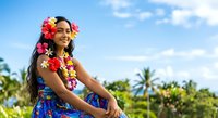 Young woman with long dark hair and South Asian ethnicity wearing colorful flower lei and floral dress, smiling outdoors in tropical setting with palm trees and blue sky for travel, vacation, and summer lifestyle content.
