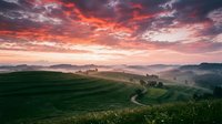 Aerial view of rolling green hills and winding rural road at sunrise with dramatic orange and pink sky, mist in valley, countryside landscape for travel and nature projects.
