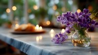Purple wildflowers in small glass vase on marble table with soft bokeh lights and candles creating romantic atmosphere for special occasion dinner setting.