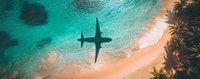 Aerial view of airplane shadow over turquoise ocean water and sandy tropical beach with palm trees, captured from above in panoramic composition.