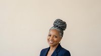 Mature African American woman with gray braided updo hairstyle wearing navy blazer, smiling against neutral background. Professional portrait for business, corporate, and lifestyle use.