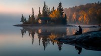 Tranquil autumn morning with person silhouetted on rocky shore, watching cabin on small island surrounded by golden trees and misty lake reflections.