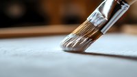 Close-up macro shot of paintbrush with natural bristles touching white paper surface, showing artistic detail and texture in warm ambient lighting.