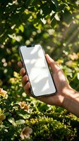 Hand holding smartphone with blank white screen outdoors in natural sunlight with green foliage background for app mockup and digital marketing presentations.
