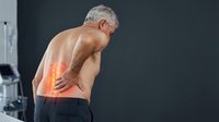 Shirtless senior Caucasian man holding lower back with glowing red pain highlight, standing in medical clinic interior.