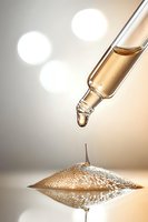 Glass dropper with golden serum drop falling onto reflective surface creating symmetrical splash pattern against soft bokeh background. Macro beauty shot.