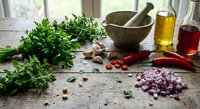 Fresh herbs parsley cilantro with red chili peppers garlic onion olive oil vinegar mortar pestle on rustic wooden kitchen counter for cooking preparation.