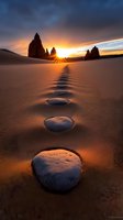 Snow-covered stepping stones lead to dramatic rock formations silhouetted against vibrant sunset sky, creating path through sandy landscape at golden hour.