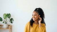 Young woman with curly dark hair and medium skin tone wearing a bright yellow shirt, smiling while touching her ear against a white wall with a potted plant, suitable for lifestyle and communication content.