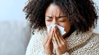 Young African American woman with curly hair sneezing into tissue, wearing cozy sweater, cold flu symptoms, healthcare concept, winter illness prevention.