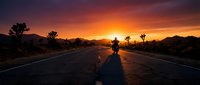 Silhouette of a person standing on an empty desert highway at sunset with vibrant orange and purple sky, mountains in distance, and sparse vegetation. Suitable for travel, adventure, and motivational content.