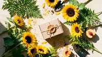 Kraft gift box surrounded by bright sunflowers, green fern leaves, and a peach on a light beige surface, flat lay composition.