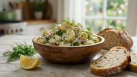 Creamy potato salad with fresh dill and herbs served in wooden bowl alongside sliced bread, perfect for lunch, picnic, or catering food photography.