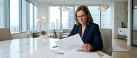 Businesswoman with brown hair and glasses wearing dark blazer reviewing documents at modern office desk with city skyline view through windows, suitable for corporate and professional business concepts.