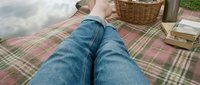 Person in jeans relaxing on checkered picnic blanket outdoors with wicker basket and books for leisure weekend activities and outdoor dining experiences.