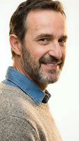 Middle-aged man with dark beard and facial hair wearing denim shirt over tan sweater, smiling at camera against white background for portrait use.