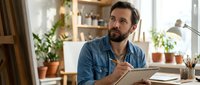 Thoughtful bearded man writing in notebook at home office desk with plants and warm lighting for creative work and planning.