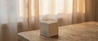 Small white gift box with ribbon on wooden table in warm sunlight streaming through window curtains creating cozy home interior atmosphere.