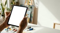 Close-up of hands holding tablet with blank white screen at desk with plants and art supplies, suitable for app mockups and digital marketing.