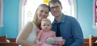 Young Caucasian family with blonde mother, brown-haired father wearing glasses, and toddler daughter in pink dress posing together in bright blue interior space for portraits, lifestyle, family bonding, parenting, and relationship content.