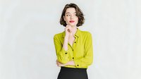 Young Caucasian woman with short brown hair wearing bright lime green shirt and black pants, thoughtfully posing against white background.