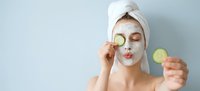 Young woman applying cucumber slices on face with clay mask for skincare routine and spa treatment at home relaxation.