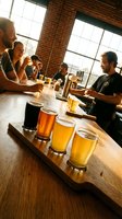 Group of men tasting craft beers at wooden table in industrial loft with brick walls and large windows, sampling flight of golden and dark ales for brewery tour or tasting event.