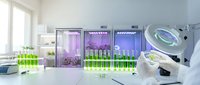 Modern laboratory interior with scientist examining plant samples under magnifying glass while green seedlings grow in illuminated cultivation chambers.