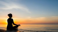 Woman silhouette meditating in lotus pose on beach at sunrise with golden sky and calm ocean waves for wellness and mindfulness concepts.