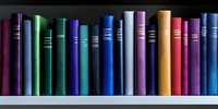 Colorful hardcover books arranged in row on white shelf, spines showing vibrant shades of purple, green, blue, pink and yellow creating rainbow effect in library.