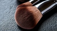 Soft makeup brush with dense synthetic bristles in rose gold color against dark textured background, dramatic side lighting creates professional cosmetic atmosphere.