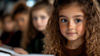 Young girl with natural curly brown hair and bright eyes wearing brown sweater smiles warmly at camera while other students work in background of classroom setting.