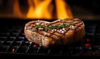 Juicy grilled beef steak with herbs and sea salt on black grill grate with fire flames in background, close up view of medium rare meat cooking.
