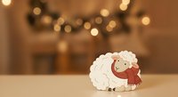 Cute sheep character wearing red scarf standing on wooden surface with warm bokeh lights and garland in blurred background, suitable for holiday greeting cards.