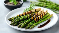 Grilled green asparagus spears with balsamic reduction served on white plate, fresh herbs and sauce in background. Perfect for restaurant menu or cooking blog.