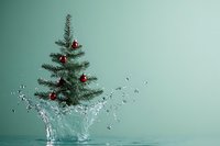 Small Christmas tree with red ornaments splashing into clear water against mint green background creating dynamic holiday concept with copy space.