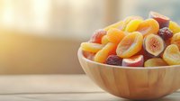Colorful assortment of dried fruits including apricots, figs, and peaches in wooden bowl on rustic table with soft natural lighting and blurred background.