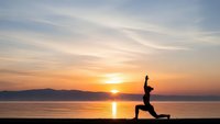 Woman practicing yoga warrior pose silhouette at sunrise by calm lake with mountain backdrop. Peaceful morning meditation and fitness routine outdoors.