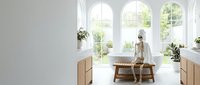 Woman in beige sweater and hat sitting on wooden bench in bright sunlit room with arched windows, potted plants, and natural light for interior design and lifestyle.